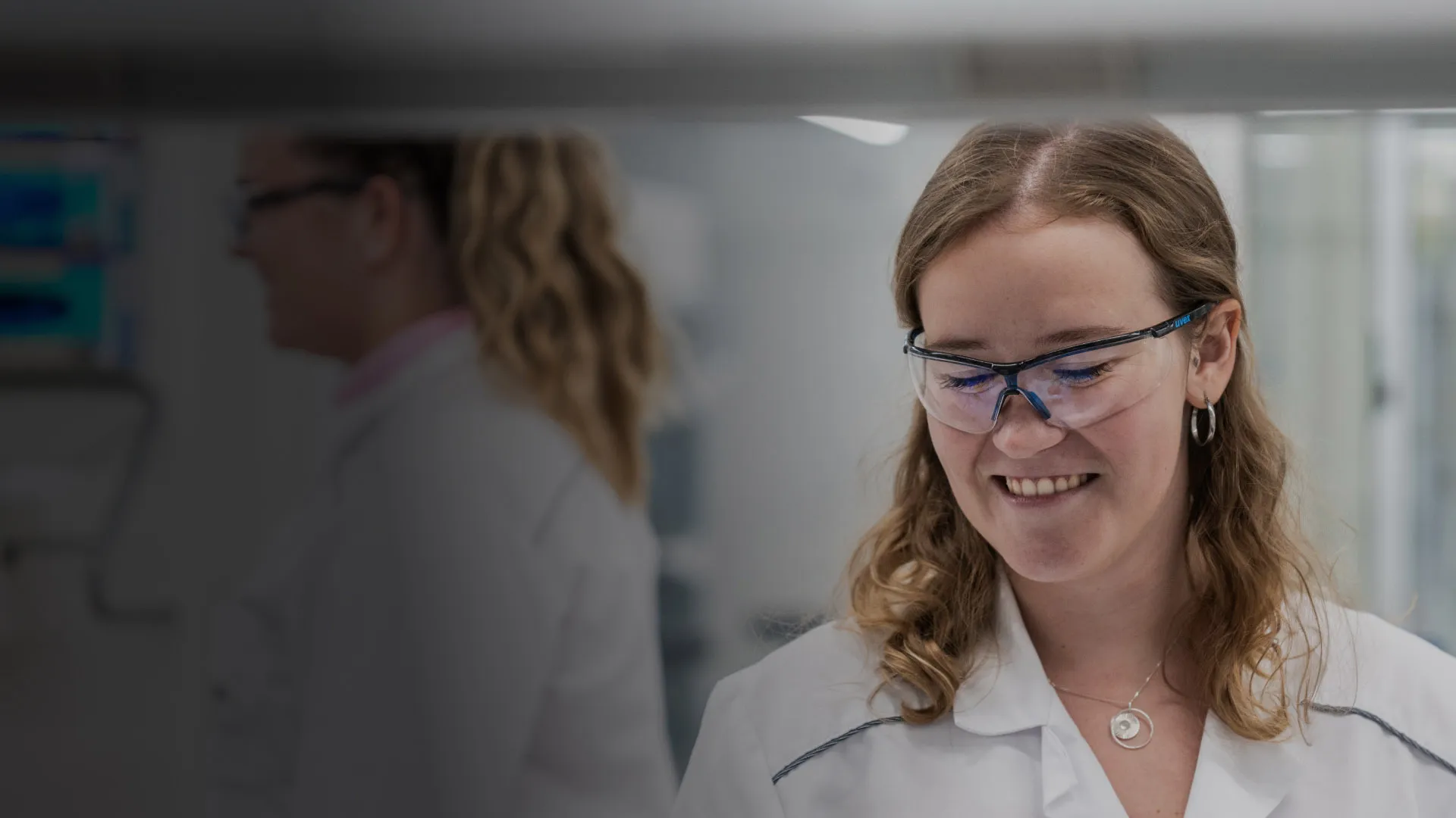 A woman in a lab coat and safety specs smiles to a colleague