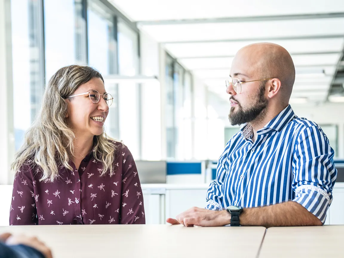 A Woman in a Patterned Maroon Shirt and a Man in a Blue and White Striped Shirt Having a Friendly Conversation Across a Table in an Office Setting.