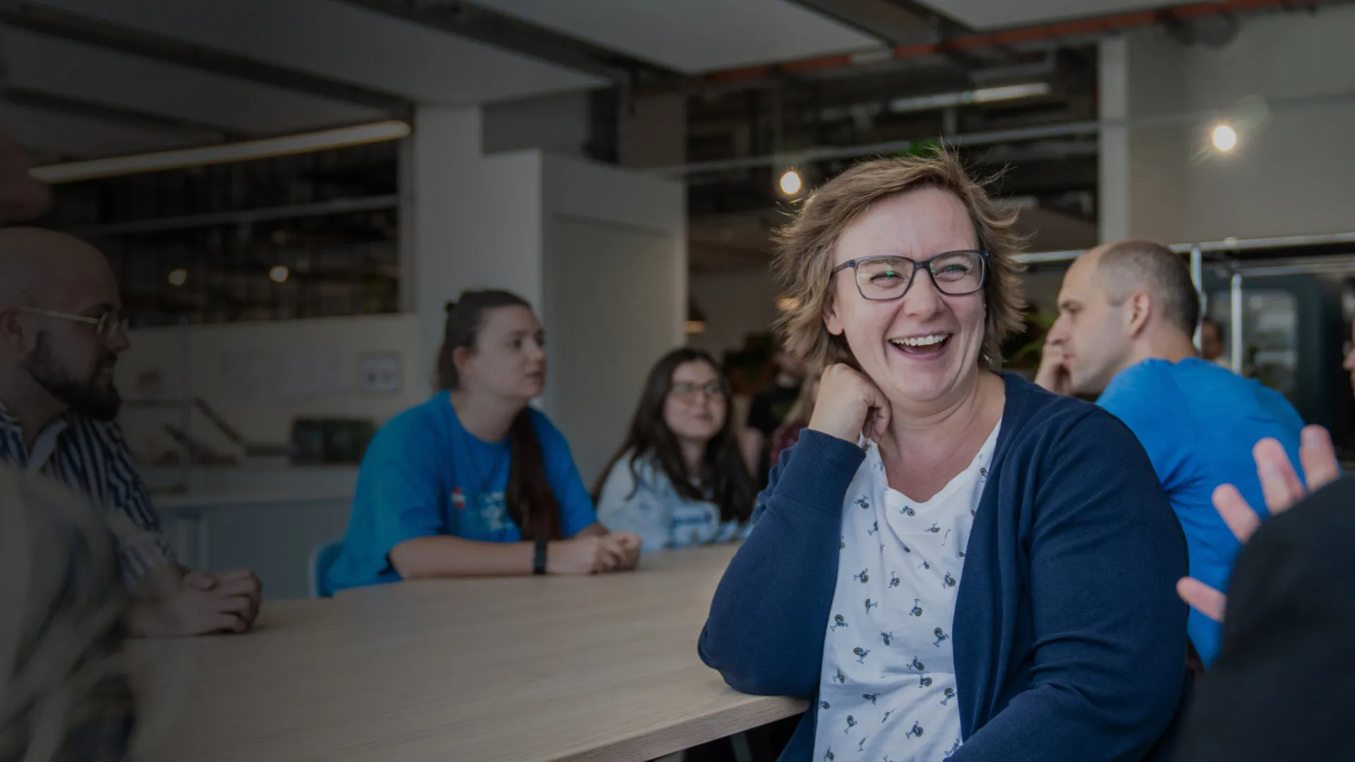 A woman in a canteen setting laughs to camera as colleagues chat around her