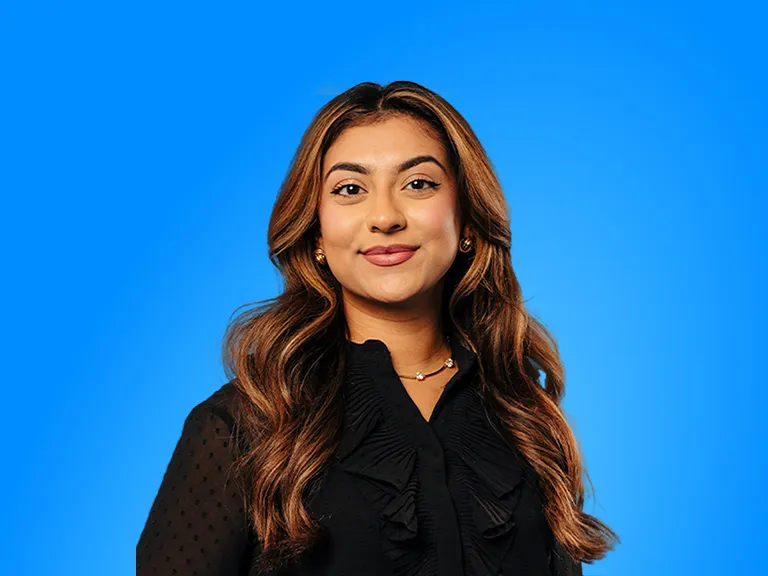 A Young Woman With Long Wavy Brown Hair Smiling Against a Solid Blue Background, Wearing a Black Ruffled Top and a Delicate Gold Necklace.