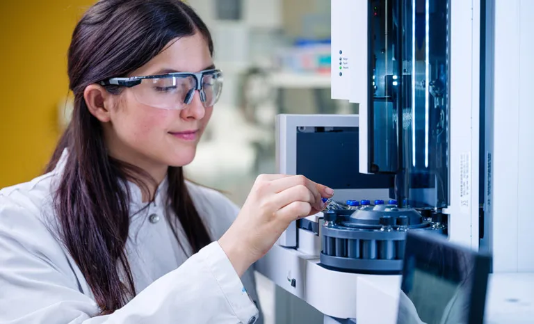 A female technician wearing protective goggles inspects a sample in a lab