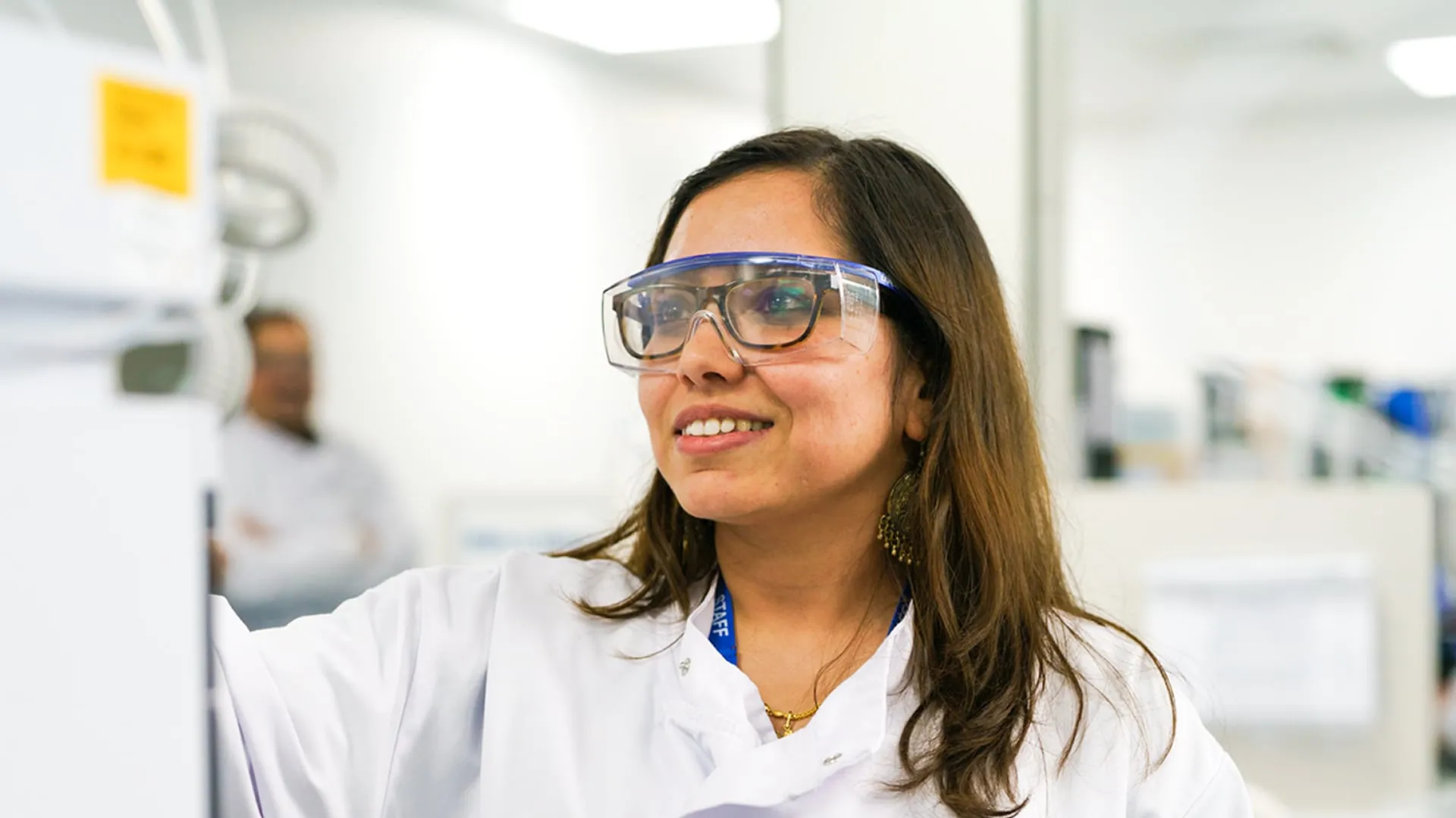 A Smiling Female Researcher Wearing a White Lab Coat and Protective Safety Goggles Works in a Bright Laboratory