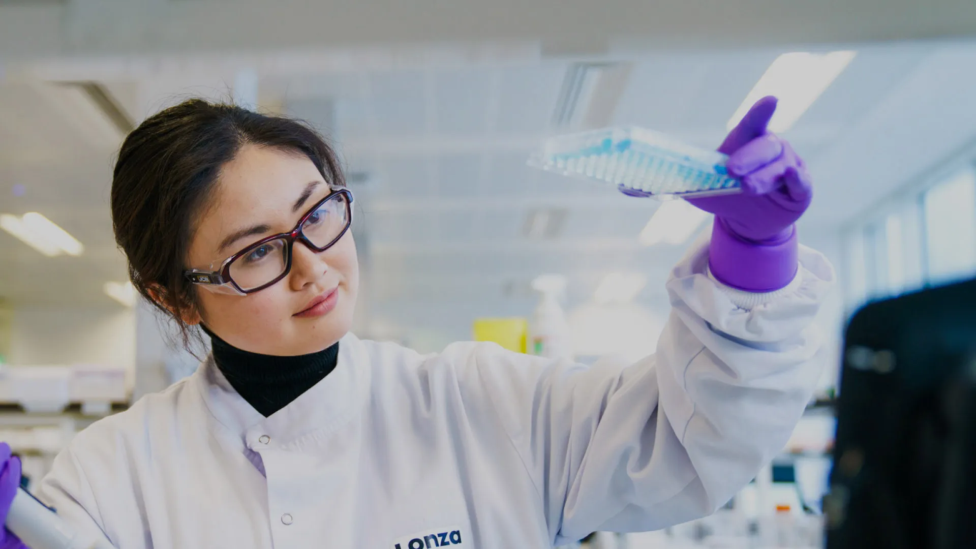 A woman in a lab coat and safety glasses inspects blue coloured samples