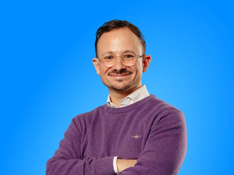 A Man With Round Glasses and a Slight Smile Posing With His Arms Crossed Against a Solid Blue Background, Wearing a Purple Sweater Over a White Collared Shi