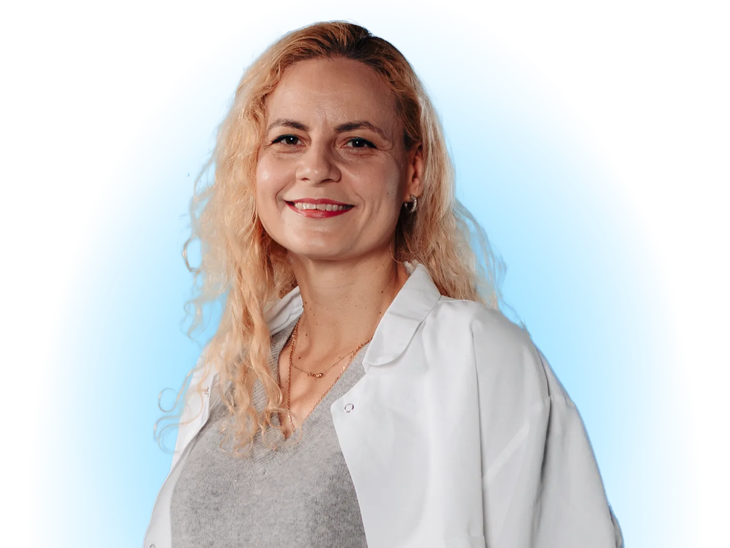 A Smiling Woman With Wavy Blonde Hair, Wearing a White Lab Coat and a Grey Sweater, Posing Against a Solid Blue Background.