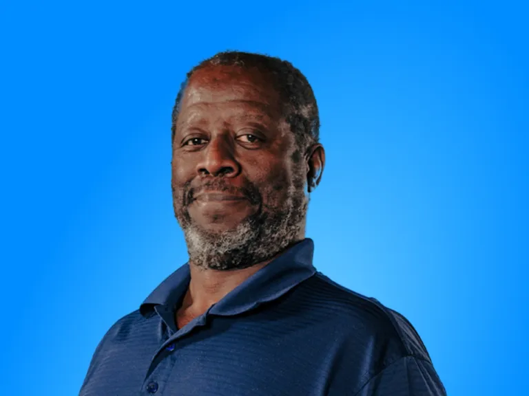 An Man With a Graying Beard Smiling Gently Against a Solid Blue Background, Wearing a Dark Blue Polo Shirt.