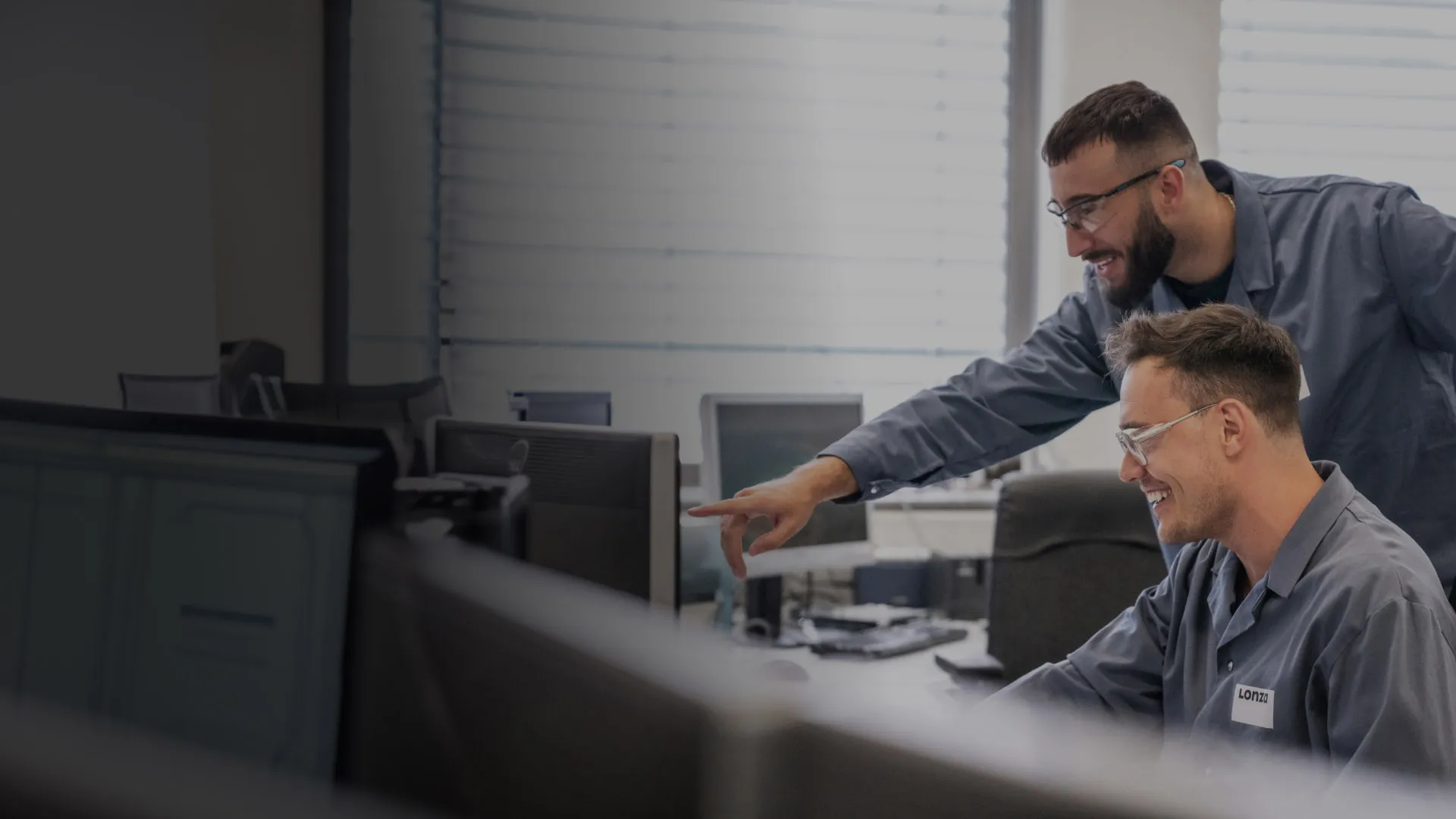 A colleague smiles as another colleague points over his shoulder to his screen