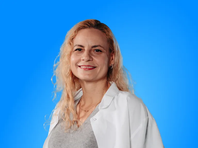 A Smiling Woman With Wavy Blonde Hair, Wearing a White Lab Coat and a Grey Sweater, Posing Against a Solid Blue Background.
