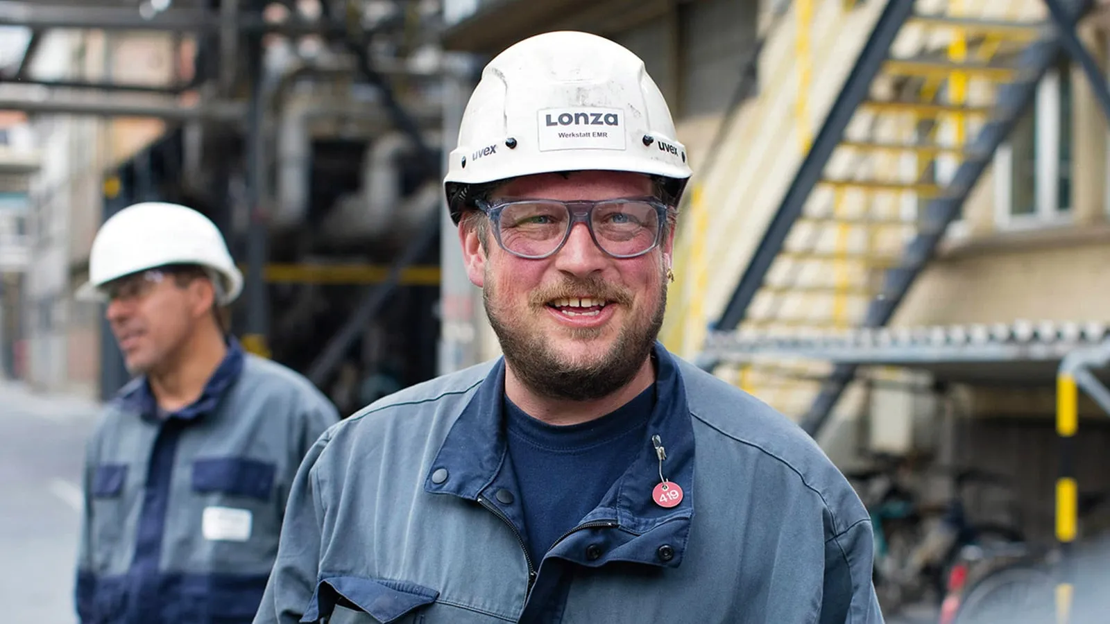 Male Engineer With Hard Hat and Safety Goggles Smiling With Colleague in Background