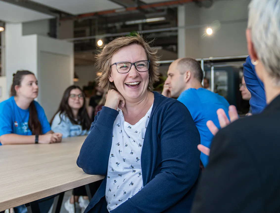 A Woman in a Canteen Setting Laughs to Camera as Colleagues Chat Around Her
