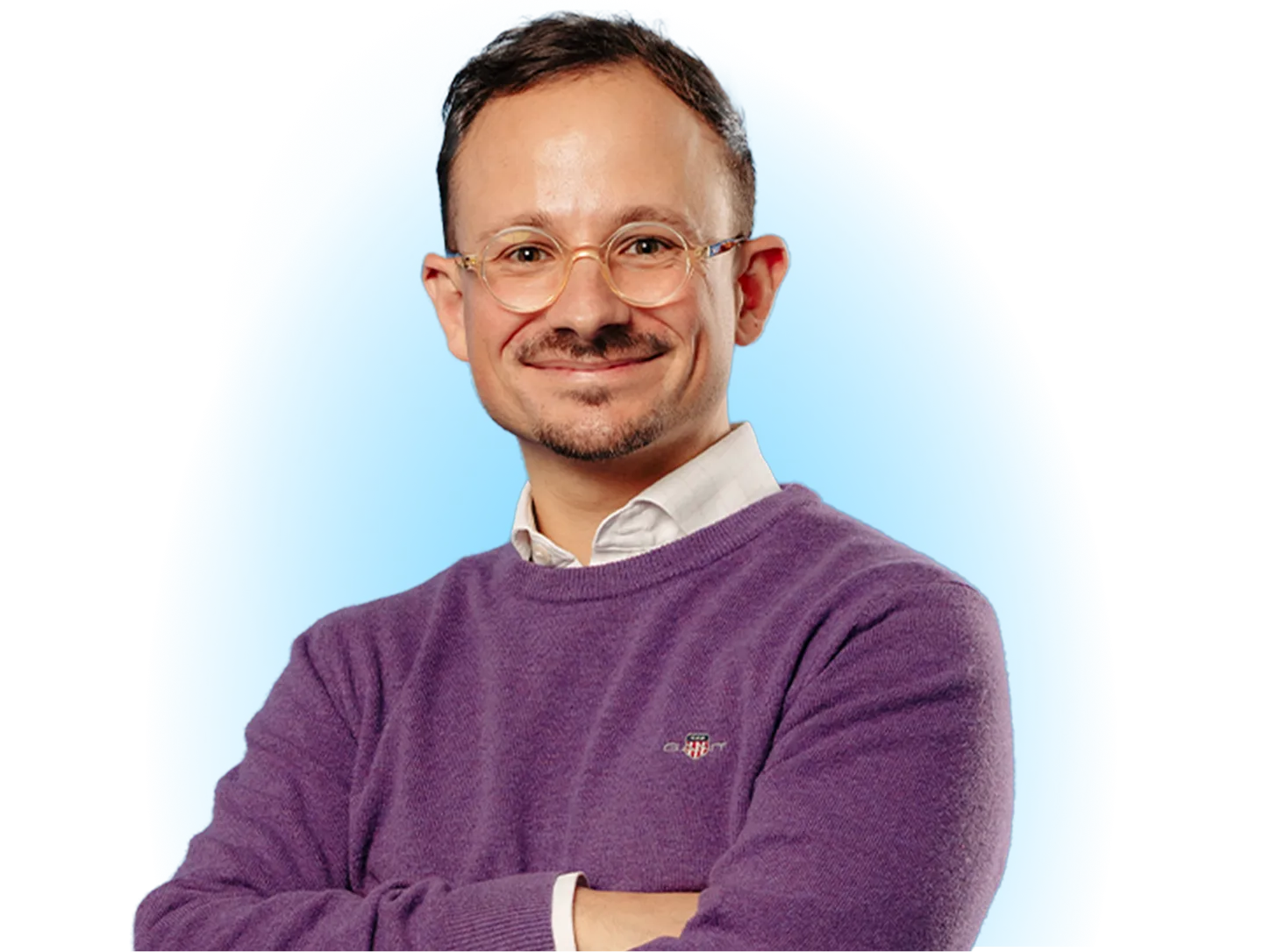 A Man With Round Glasses and a Slight Smile Posing With His Arms Crossed Against a Solid Blue Background, Wearing a Purple Sweater Over a White Collared Shi (1)